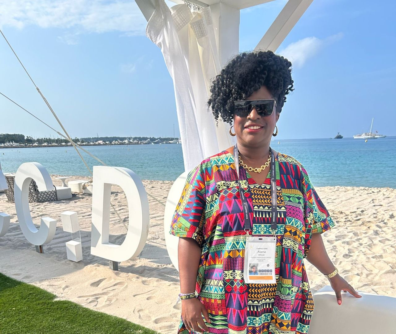 A woman poses on a sunny beach in front of large white letters wearing a vibrant, multi-colored geometric print dress with long black fringe at the bottom. She accessorizes with sunglasses, gold jewelry, a LinkedIn event badge, and cream sneakers, standing confidently near white lounge seating with the ocean and yachts in the background.